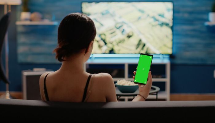 The image shows a woman holding a phone, sitting in front of the TV. — Freepik