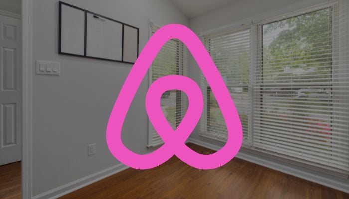 The image shows the logo of Airbnb with a room in the background. — Canva