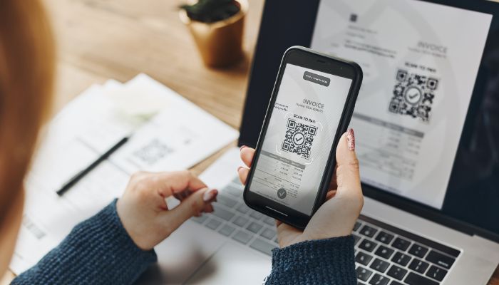 An undated image of a person scanning QR code on a phone through the laptop screen. — Canva